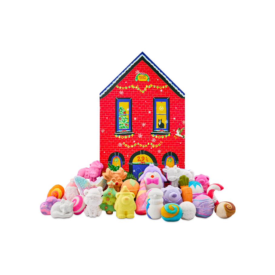 A red brick house-shaped gift box stands behind a large display of festive bath products, including a sleeping fox, a bear, a penguin, and a Christmas tree bath bomb, among many more themed products.